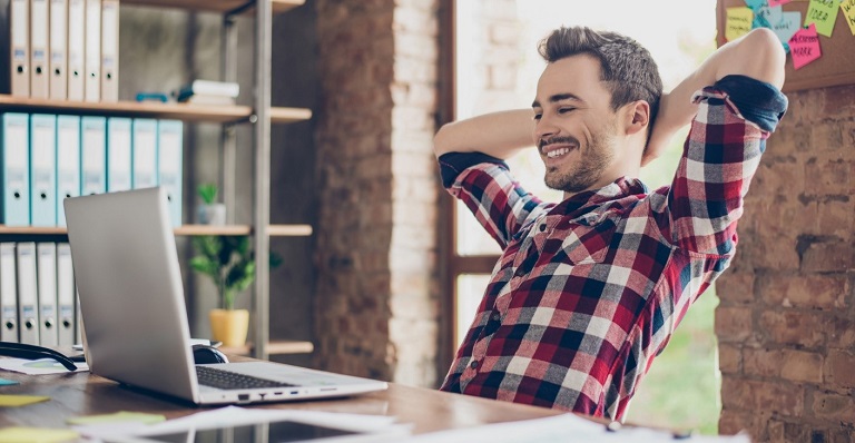 Cheerful young brunet guy at his work place staring at his laptop screen