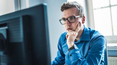 Un jeune homme d'affaires portant des lunettes est assis à un bureau et se concentre sur l'écran de son ordinateur.