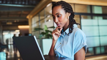 Thoughtful young Black professional with braided hair studies laptop, chin resting on hand, in open-plan office