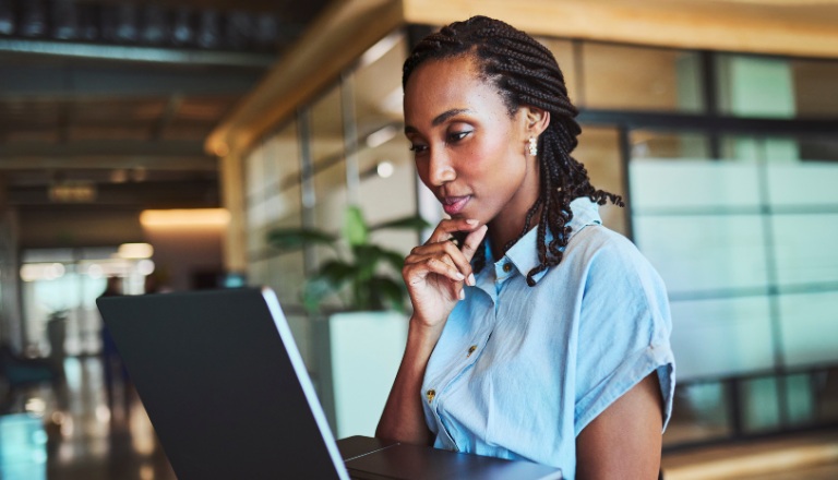 Thoughtful young Black professional with braided hair studies laptop, chin resting on hand, in open-plan office