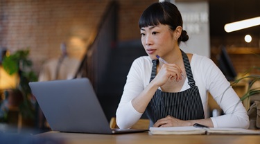 Upscale coffee shop owner wearing apron sits thoughtfully with laptop and notebook, planning menu.