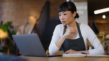 Upscale coffee shop owner wearing apron sits thoughtfully with laptop and notebook, planning menu.