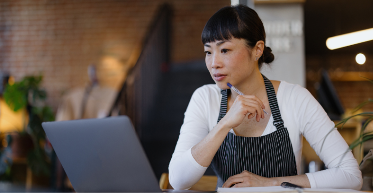 Upscale coffee shop owner wearing apron sits thoughtfully with laptop and notebook, planning menu.