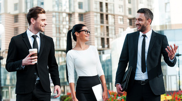 Trois professionnels en tenue de ville discutent en marchant dans une place publique moderne, café et tablette à la main.