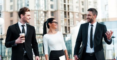 Three business professionals in suits walk and talk through a modern city plaza, holding coffee and a tablet.