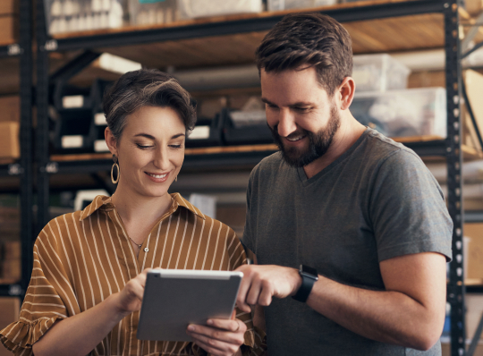 Two people reviewing inventory and supply chain information using a tablet in a warehouse.