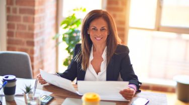 Une femme souriante, assise sur une chaise, lit des documents à son bureau.