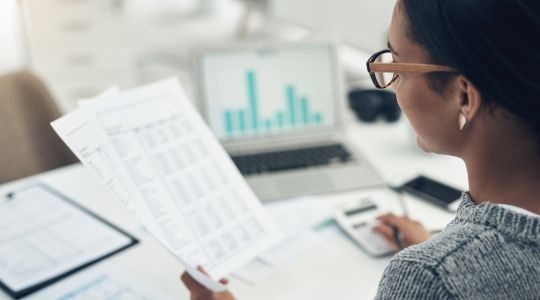 Une femme avec des lunettes regarde des documents fiscaux, sur papier et à l’ordinateur.