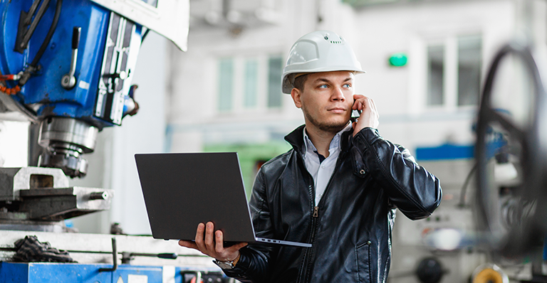 An engineer standing in front of an industrial machine with his laptop talks on the phone