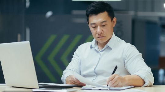 A businessman sitting at a desk, signing papers with a pen, with a laptop in front of him.