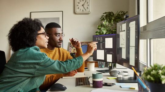 A man and a woman sitting at a desk, looking at charts on two monitors, with the woman pointing at the screens with a pen.