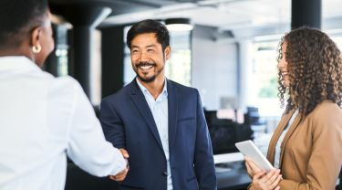 Trois professionnels participent à une réunion d’affaires ou à une discussion dans un bureau moderne. Deux personnes, dont une en costume bleu, se serrent la main, alors qu’une troisième se tient à proximité, une tablette à la main. L’arrière-plan présente de grandes fenêtres et un ameublement de bureau contemporain qui créent  une atmosphère lumineuse et collaborative.