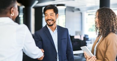 Trois professionnels participent à une réunion d’affaires ou à une discussion dans un bureau moderne. Deux personnes, dont une en costume bleu, se serrent la main, alors qu’une troisième se tient à proximité, une tablette à la main. L’arrière-plan présente de grandes fenêtres et un ameublement de bureau contemporain qui créent  une atmosphère lumineuse et collaborative.