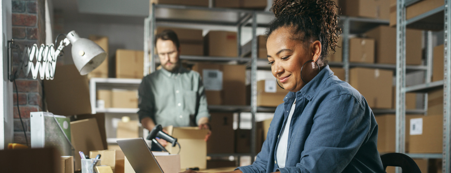 Two warehouse employees: one using a laptop while the other checks boxes in a room full of cardboard boxes on shelves.