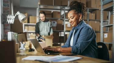  Two warehouse employees: one using a laptop while the other checks boxes in a room full of cardboard boxes on shelves.