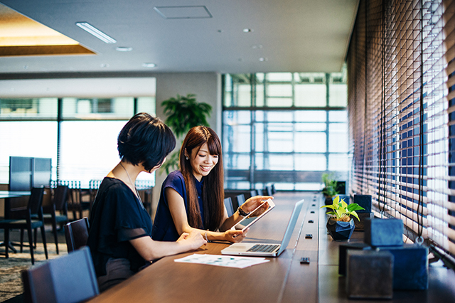 Two Japanese businesswomen smiling and collaborating while looking at a tablet in a coffee shop.