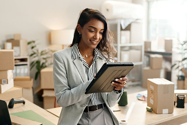 A smiling Indian woman using a tablet to oversee her business operations, surrounded by boxes and packages, representing the logistics of an e-commerce business.