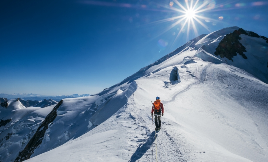 Un alpiniste en solo, vêtu d’une veste orange, marchant sur une étroite crête enneigée sous un soleil radieux, avec de majestueux sommets alpins au loin