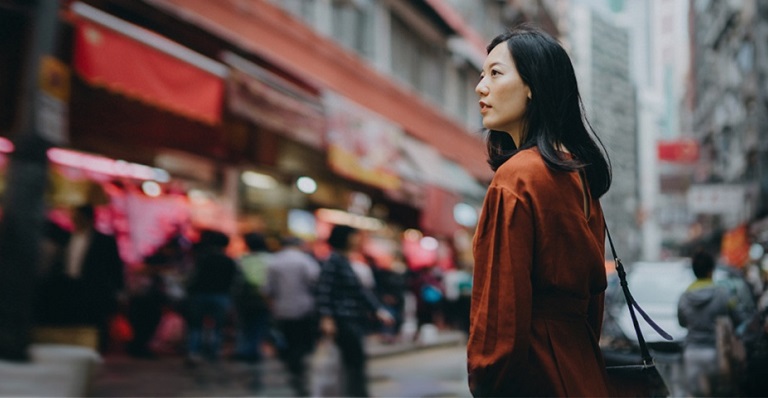 A woman stands on a busy street, surrounded by tall buildings and looking pensive.