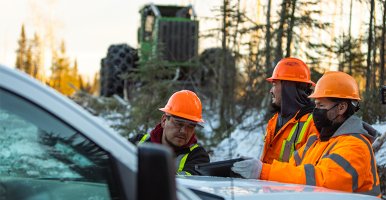 Three men, wearing hard hats and vests, stand beside a vehicle looking at a tablet together. Behind them is a green tractor.