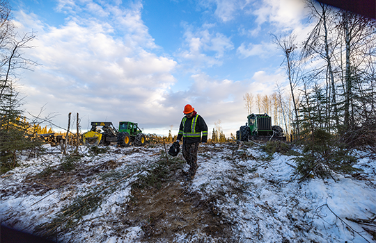 A man in a hard hat and vest is walking in a field, with tractors behind him clearing trees.