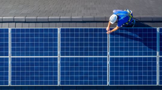 A worker in safety gear installs solar panels on a rooftop.