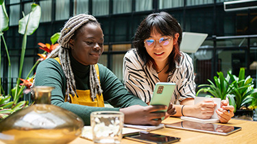 Image of two women with mobile phone, notepad and tablets flourishing in an inclusive, equitable workspace.