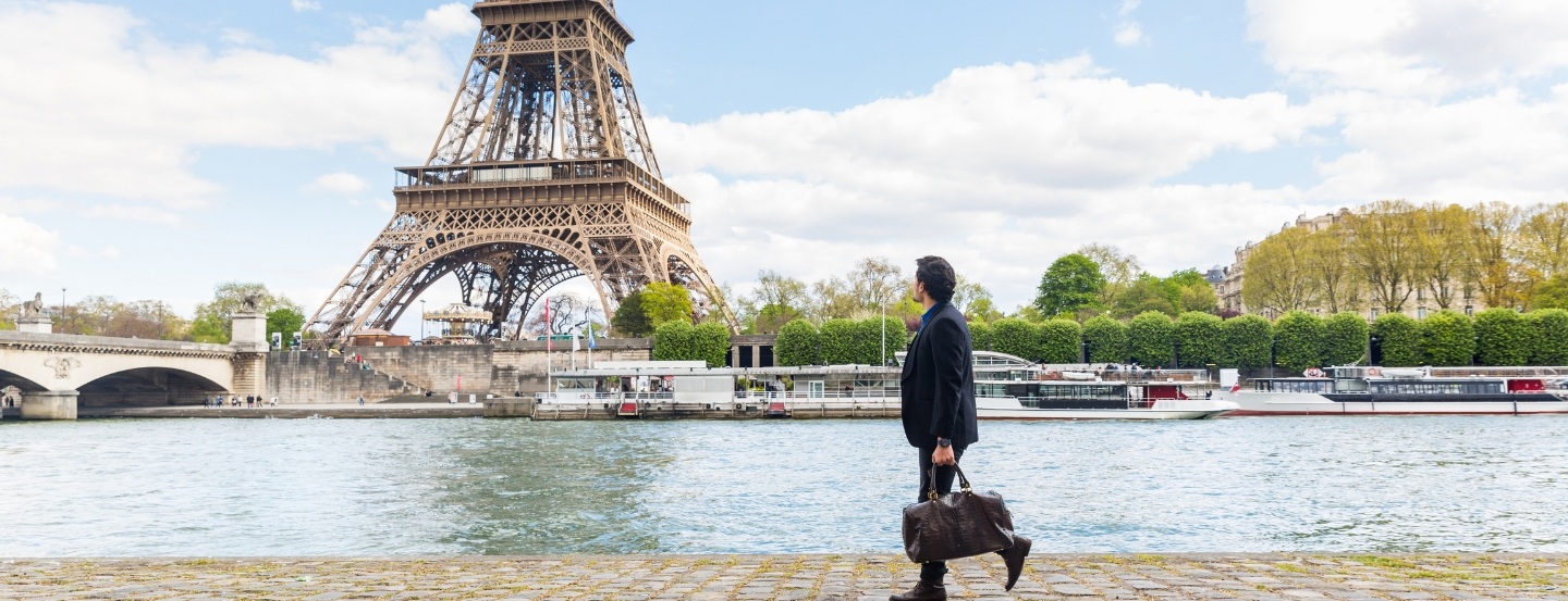 Une personne portant un sac de voyage qui marche le long d’une promenade au bord de la rivière, avec la Tour Eiffel et des bateaux de croisière en arrière-plan.