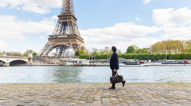 Person carrying a travel bag while walking along a riverside walkway, with the Eiffel Tower and riverboats in the background.