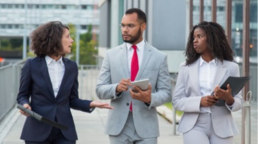 Three business professionals walking and conversing while walking along a modern building.