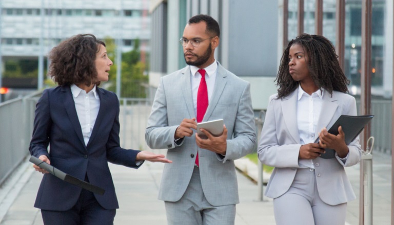 Three business professionals walking and conversing while walking along a modern building.