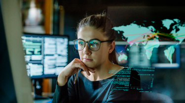 Jeune femme, portant des lunettes, entourée d'écrans d'ordinateur dans un bureau sombre, avec un écran montrant une carte du monde.