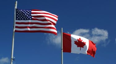 U.S. and Canadian flags wave in the wind with a blue sky and clouds in the background.