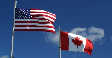 U.S. and Canadian flags wave in the wind with a blue sky and clouds in the background.