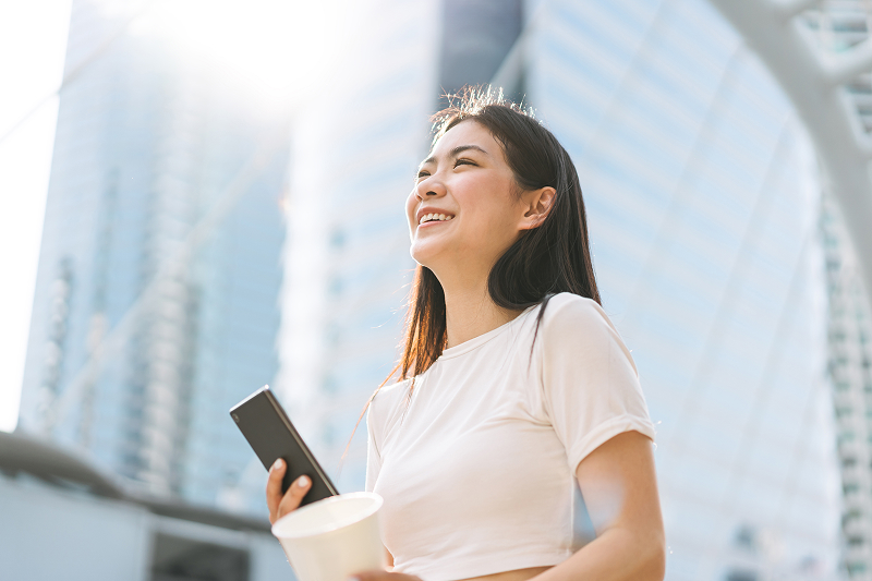 Person outdoors holding a smartphone and coffee cup, smiling in front of city buildings.