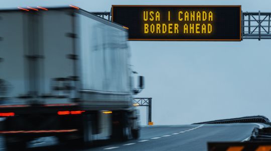 Blurry semi-truck approaching a highway sign for the U.S.-Canada border.
