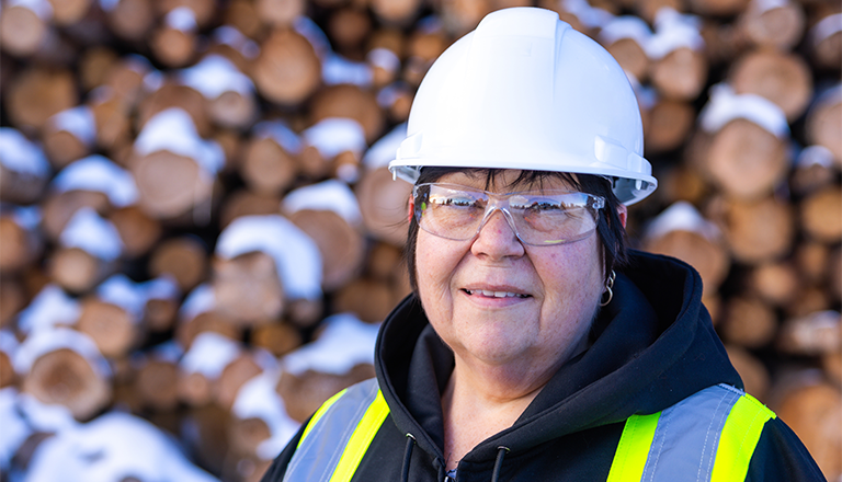 Female industrial worker with white hard hat and yellow safety vest standing in front of a blurry wood pile.