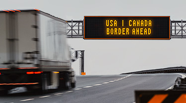 Transport truck passes under USA-Canada Border ahead sign on highway.