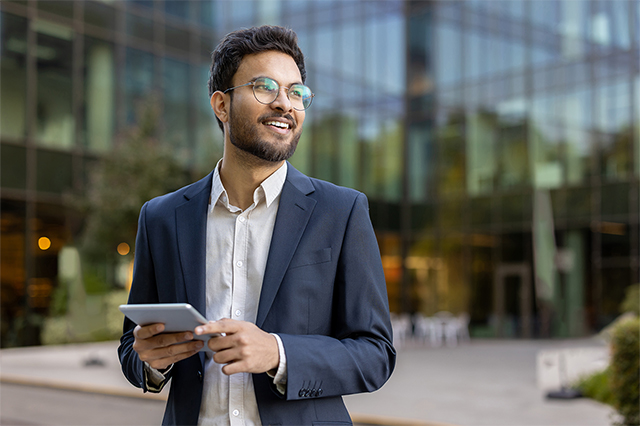 Confident businessman in glasses and suit using a tablet outdoors with a modern office building in the background, looking outward. Represents professional success, technology, and business lifestyle. 