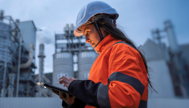 Woman wearing a hard hat using a tablet at an industrial facility, reviewing operations and supply chain information in a manufacturing environment.