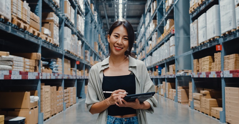 Person standing in a warehouse full of boxes holding a tablet and a pencil