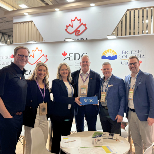Un groupe de personnes debout côte à côte et souriant devant un stand de salon professionnel où sont visibles les logos d’EDC, du Canada et de la Colombie Britannique.